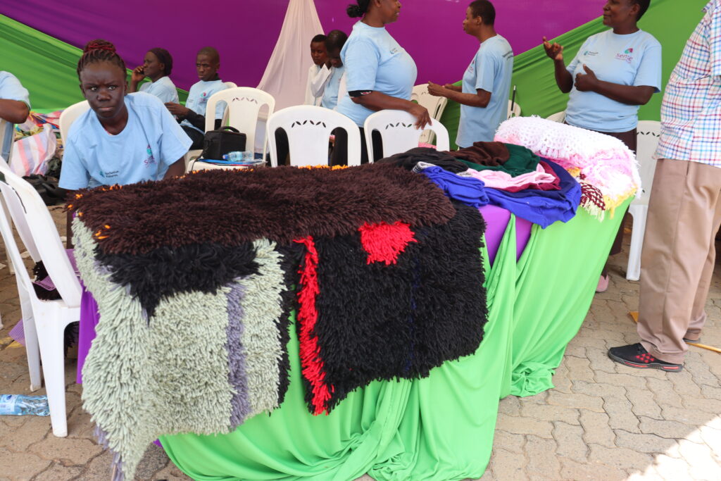 A young adult Kenyan woman sits at a table where she sells handcrafted carpets