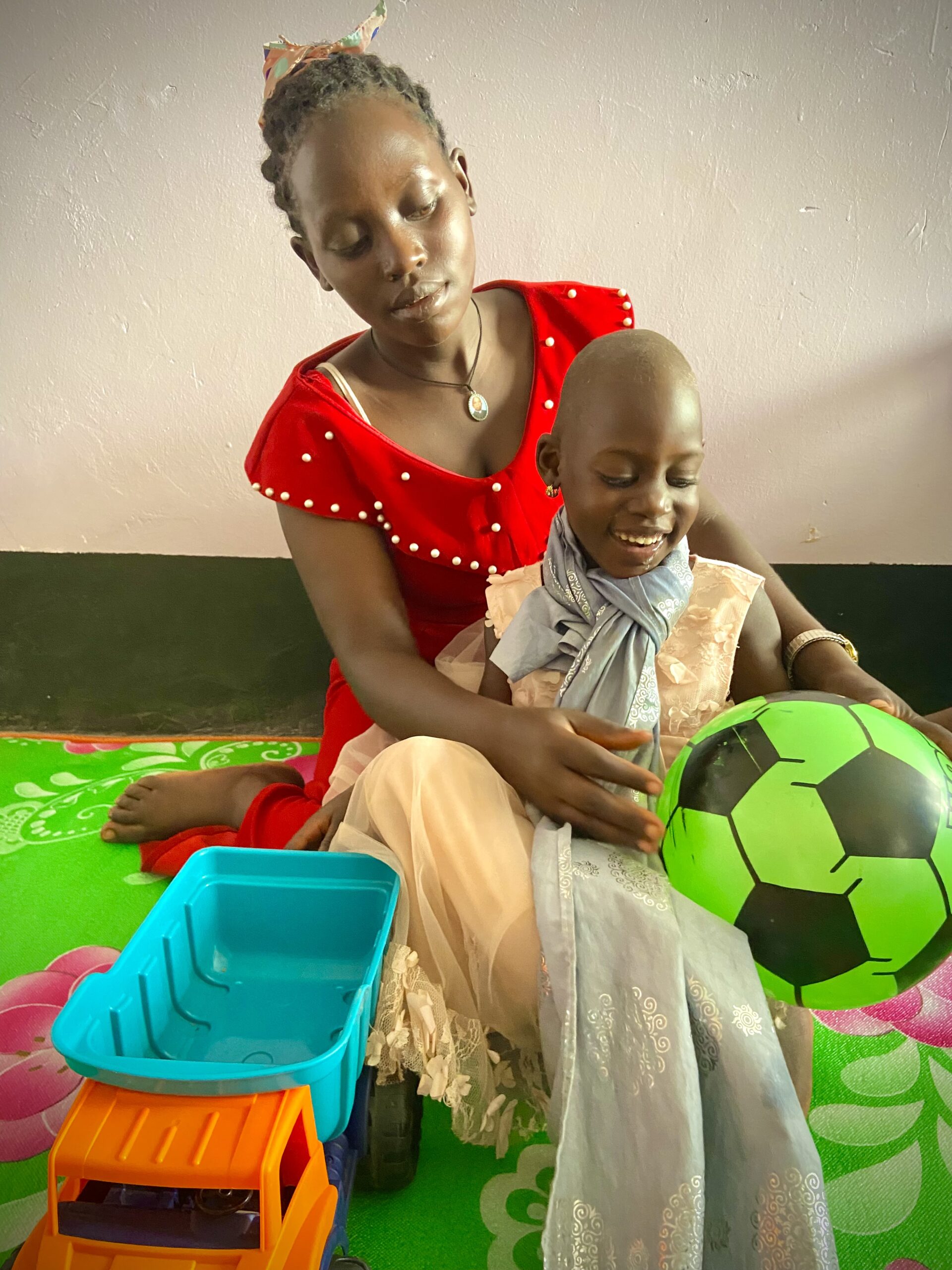 A young Ugandan mother wearing a red dress plays with her small daughter and a big green ball