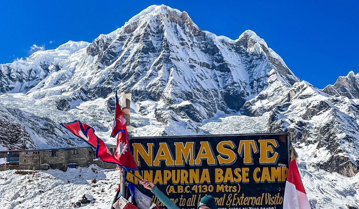 The snowy white mountain peaks of Annapurna loom in the background. In the foreground, a large dark green banner reads 'Namaste Annapurna Base Camp'
