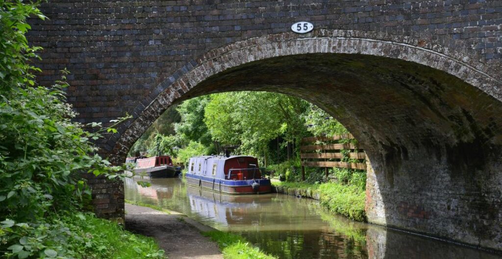 An old grey stone bridge surrounded by greenery over a canal with parked canal boats