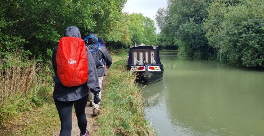 A line of hikers in coats and backpacks trek alongside a canal with a parked canal boat