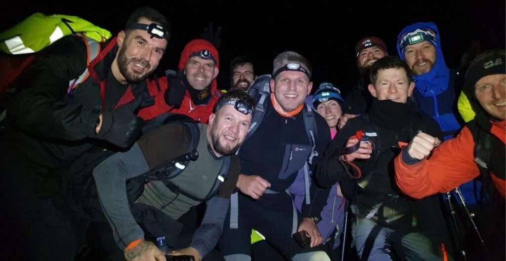 A big group of hikers wearing climbing gear and head torches gather for a selfie in the middle of the night