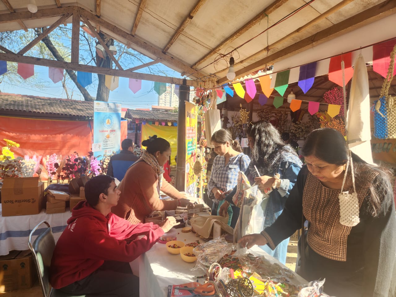 A market stall on a sunny day decorated with brightly coloured flags. A young Indian man with deafblindness greets his customers.