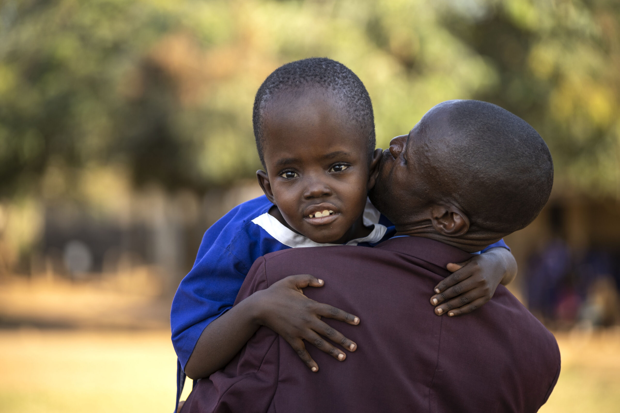 A Ugandan man wearing a dark purple suit with his back to the camera holds a young girl wearing school uniform, who is looking at the camera, and kiss her cheek.