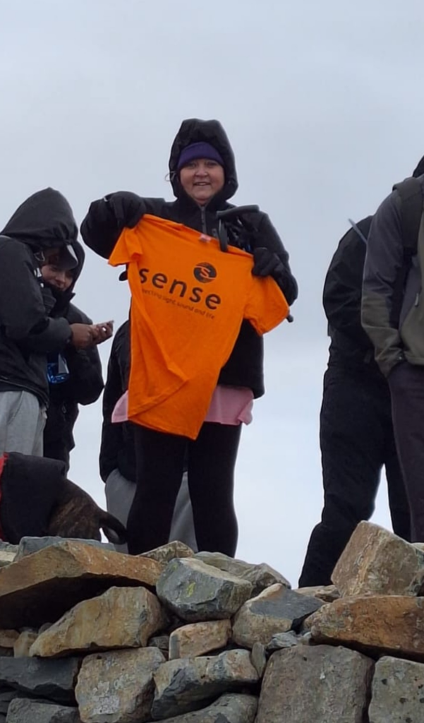 A woman in hiking gear and a woolly hat holds up a neon orange Sense branded t-shirt.