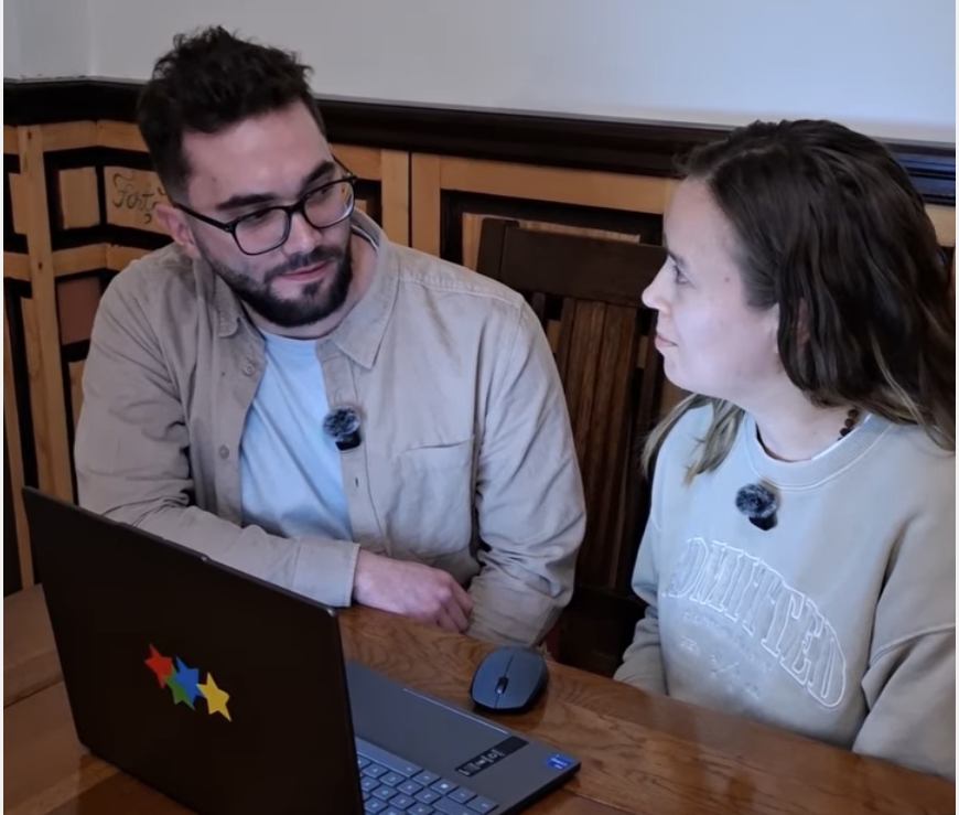 A young adult Romanian man with dark hair and glasses and woman with long brown hair talk to one another whilst using a laptop