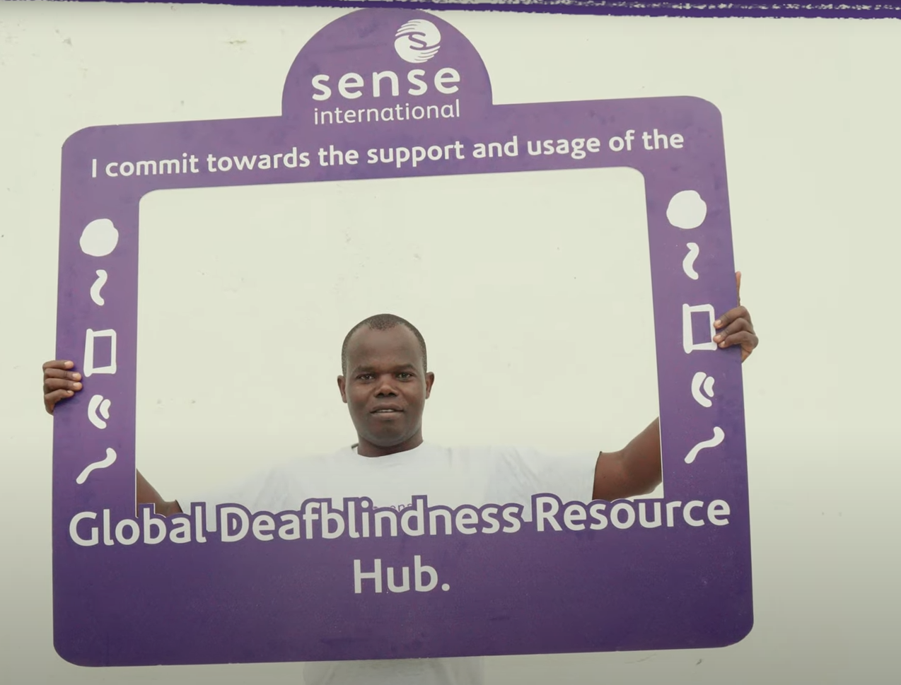 An adult Kenyan man holds up a large purple frame featuring the Sense International logo