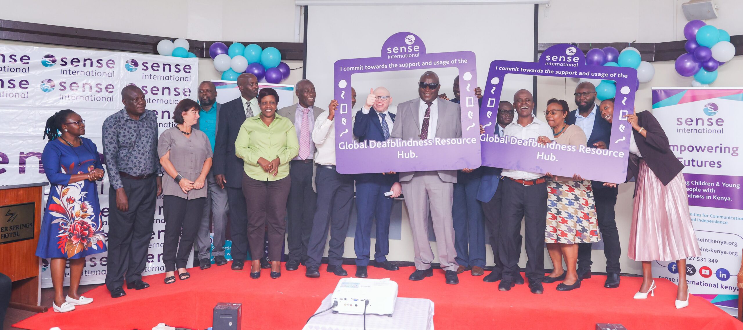 A large group of Kenyan adults stand on a red stage surrounded by Sense International logos and balloons. They hold large purple signs for the Global Deafblindness Resource Hub.