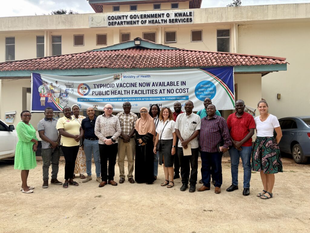 A large group of Kenyan adults and two British visitors stand in front of a banner proclaiming that the typhoid vaccine is now freely available.