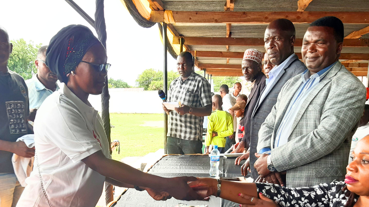 A Tanzanian woman wearing a headscarf and glasses shakes hands over a table